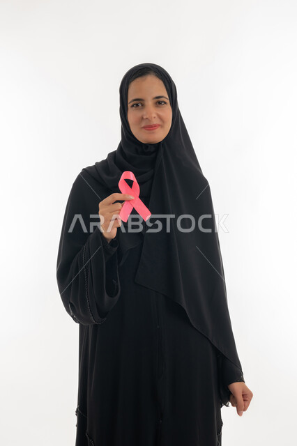 رمز الحملة العالمية لسرطان الثدي، تحدي الأمراض الخبيثة، بورتريه لسيده عربية خليجية إماراتية ترتدي الحجاب والعباءة السوداء تحمل بيديها الشريط الوردي، شابة  تنظر الى الكاميرا بإيماءات البهجة والتفاؤل، حملة نسائية توعوية للكشف المبكر عن سرطان الثدي، خلفية بيضاء