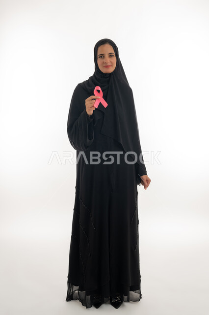 Breast cancer global campaign symbol, challenge malignant diseases, portrait of an Emirati Gulf Arab woman wearing a hijab and a black abaya holding a pink ribbon in her hands, a young woman looking at the camera with gestures of joy and optimism, a women's awareness campaign for early detection of breast cancer, white background