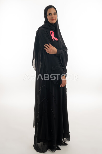 Breast cancer global campaign symbol, challenge malignant diseases, portrait of an Arab Gulf Emirati woman wearing a hijab and a black abaya wearing a pink ribbon, a young woman pointing her hand with standing gestures to prevent the spread of the disease, a women's awareness campaign for early detection of breast cancer, white background