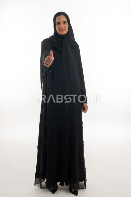 تعابير الموافقة و القبول ,النظر الى الكاميرا بإيماءات السرور، بورتريه لسيده عربيه خليجيه اماراتيه ترتدي العباءه والحجاب تشير باصبع الابهام نحو الاعلى (علامه الامتياز) بايماءات الرضى والاعجاب، خلفية بيضاء