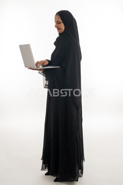تصفح الانترنت وبرامج التواصل الاجتماعي, إستخدام الحاسوب لإنجاز المهام، بورتريه لسيده عربيه خليجيه اماراتيه ترتدي العباءه والحجاب تحمل بيدها جهاز اللاب توب المحمول، تطور التكنولوجيا والأجهزة التقنية، خلفية بيضاء