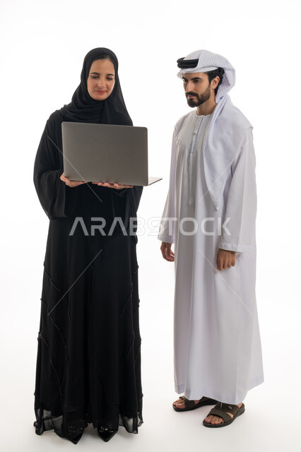 The concept of using modern technology in performing tasks and working remotely, cooperation and sharing between colleagues in choosing a work plan, a portrait of an Emirati Gulf Arab man and woman wearing traditional Emirati clothing working on a laptop to follow up on the progress of work, white background