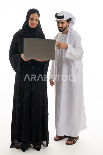 مفهوم استخدام التكنولوجيا الحديثة في أداء المهام والعمل عن بعد، التعاون والتشارك بين الزملاء في اختيار خطة العمل، بورتريه لسيده ورجل عربيان خليجيان اماراتيان يرتديان الزي الاماراتي التقليدي يقومان بالعمل على جهاز الاب توب المحمول في متابعه سير العمل، خلفيه بيضاء