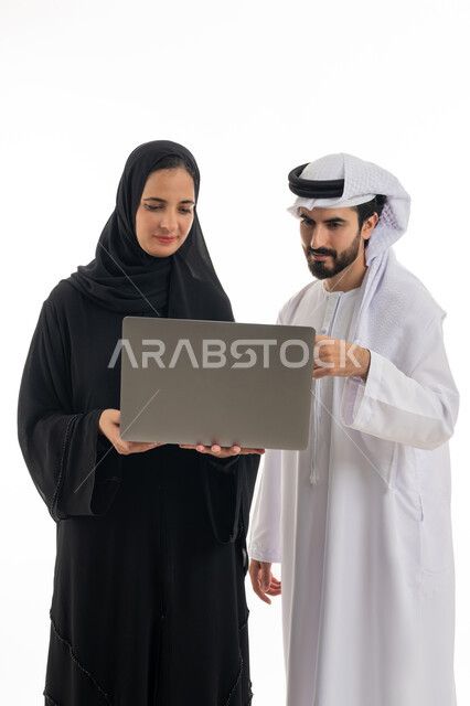 مفهوم استخدام التكنولوجيا الحديثة في أداء المهام والعمل عن بعد، التعاون والتشارك بين الزملاء في اختيار خطة العمل، بورتريه لسيده ورجل عربيان خليجيان اماراتيان يرتديان الزي الاماراتي التقليدي يقومان بالعمل على جهاز الاب توب المحمول في متابعه سير العمل، خلفيه بيضاء