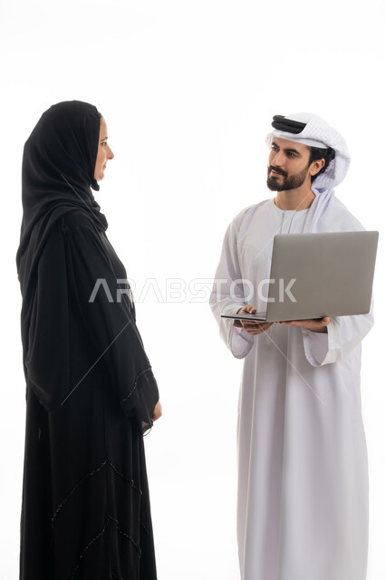 مفهوم استخدام التكنولوجيا الحديثة في أداء المهام والعمل عن بعد، التعاون والتشارك بين الزملاء في اختيار خطة العمل، بورتريه لسيده ورجل عربيان خليجيان اماراتيان يرتديان الزي الاماراتي التقليدي يقومان بالعمل على جهاز الاب توب المحمول في متابعه سير العمل، خلفيه بيضاء