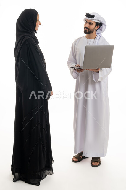 مفهوم استخدام التكنولوجيا الحديثة في أداء المهام والعمل عن بعد، التعاون والتشارك بين الزملاء في اختيار خطة العمل، بورتريه لسيده ورجل عربيان خليجيان اماراتيان يرتديان الزي الاماراتي التقليدي يقومان بالعمل على جهاز الاب توب المحمول في متابعه سير العمل، خلفيه بيضاء