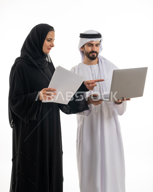 مفهوم استخدام التكنولوجيا الحديثة في أداء المهام والعمل عن بعد، التعاون والتشارك بين الزملاء في اختيار خطة العمل، بورتريه لسيده ورجل عربيان خليجيان اماراتيان يرتديان الزي الاماراتي التقليدي يقومان بالعمل على جهاز الاب توب المحمول في متابعه سير العمل، خلفيه بيضاء