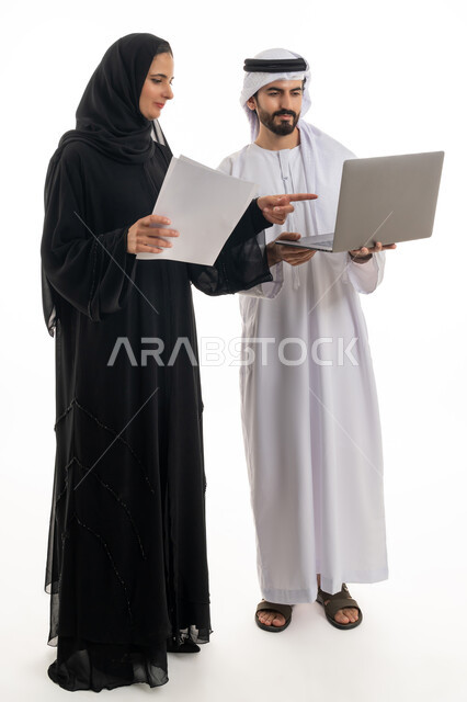 مفهوم استخدام التكنولوجيا الحديثة في أداء المهام والعمل عن بعد، التعاون والتشارك بين الزملاء في اختيار خطة العمل، بورتريه لسيده ورجل عربيان خليجيان اماراتيان يرتديان الزي الاماراتي التقليدي يقومان بالعمل على جهاز الاب توب المحمول في متابعه سير العمل، خلفيه بيضاء