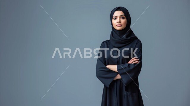 Elegance and interest in appearance, the concept of strength and self-confidence, close-up portrait of a Saudi Arabian Gulf woman wearing a hijab and a black abaya standing with crossed hands looking at the camera, gray background