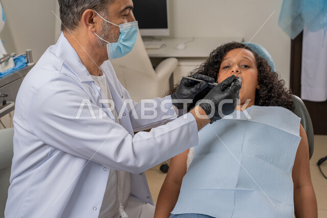 استخدام معدات وأدوات طب الأسنان، زيارة العيادات الصحية وإتخاذ الإجراءات اللازمة، الأساليب العلاجية المتبعة للعناية بالفم، معالجة التسوس وتركيب الحشوات، صورة مقربة لطبيب عربي خليجي اماراتي يرتدي القفازات والكمامة يقوم بفحص أسنان الفتاة المريضة