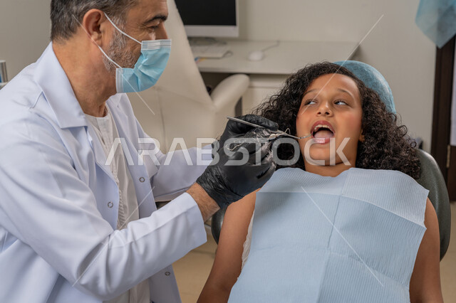 استخدام معدات وأدوات طب الأسنان، زيارة العيادات الصحية وإتخاذ الإجراءات اللازمة، الأساليب العلاجية المتبعة للعناية بالفم، معالجة التسوس وتركيب الحشوات، صورة مقربة لطبيب عربي خليجي اماراتي يرتدي القفازات والكمامة يقوم بفحص أسنان الفتاة المريضة