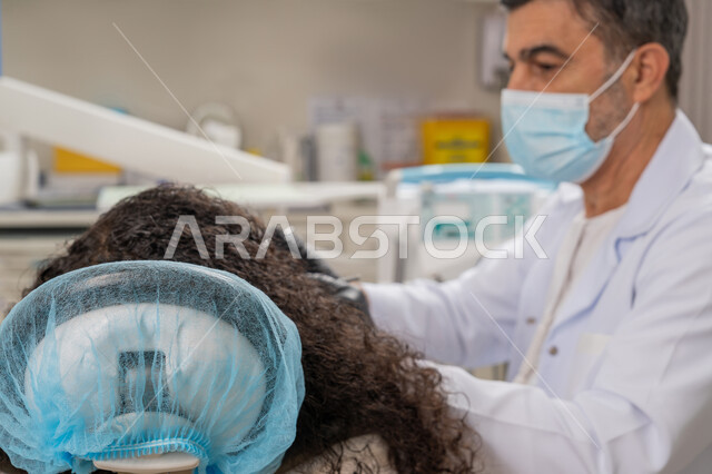 ضرورة زيارة العيادات السنية بشكل دوري، الأساليب العلاجية المتبعة للعناية بالفم، معالجة التسوس وتركيب الحشوات، صورة مقربة لطبيب عربي خليجي اماراتي يرتدي القفازات والكمامة يقوم بفحص أسنان الفتاة المريضة، استخدام معدات وأدوات طب الأسنان