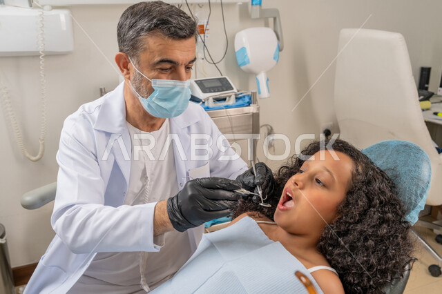 استخدام معدات وأدوات طب الأسنان، زيارة العيادات الصحية وإتخاذ الإجراءات اللازمة، الأساليب العلاجية المتبعة للعناية بالفم، معالجة التسوس وتركيب الحشوات، صورة مقربة لطبيب عربي خليجي اماراتي يرتدي القفازات والكمامة يقوم بفحص أسنان الفتاة المريضة