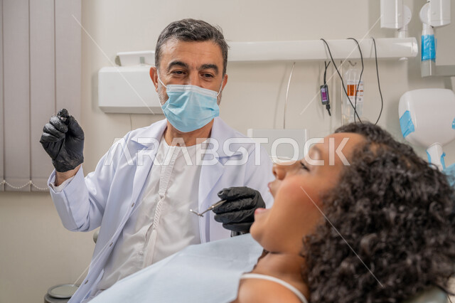 Visiting health clinics and taking the necessary measures, therapeutic methods used for oral care, treating caries and installing fillings, a close-up image of an Arab Gulf Emirati doctor wearing gloves and a mask examining the teeth of a sick girl, using dental equipment and tools