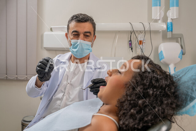 Visiting health clinics and taking the necessary measures, therapeutic methods used for oral care, treating caries and installing fillings, a close-up image of an Arab Gulf Emirati doctor wearing gloves and a mask examining the teeth of a sick girl, using dental equipment and tools