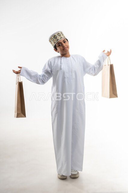 The joy of shopping in the Sultanate of Oman, taking advantage of the White Friday offers and discounts, purchasing supplies and gifts, a portrait of a smiling Omani Gulf Arab child wearing a dishdasha and a cloak, holding cardboard paper bags in his hand and raising them up, full-length body, white background