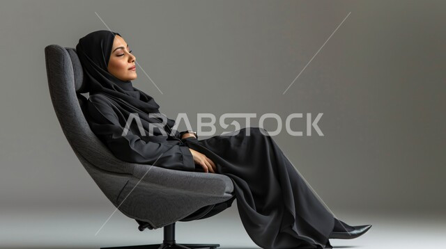  الشعور بالنعاس والحاجة الى النوم، أخذ قسط من الراحة، اغماض العينين للتأمل وشرود الذهن، بورتريه لإمرأة عربيه خليجيه سعوديه محجبه ترتدي العباءة تجلس على كرسي مريح بإيماءات الاسترخاء، خلفية رمادية