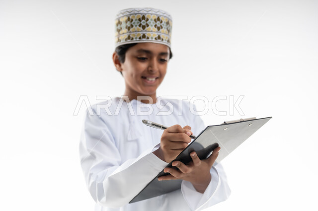 مراجعة الدروس وحل الواجبات، المناهج العلمية التعليمية، العودة الى المدارس، مفهوم الجد والاجتهاد للوصول إلى النجاح، بورتريه مقرب لطالب عربي خليجي عماني  يرتدي الدشداشة والكمة يحمل قلم وستاند ورق يقوم بكتابة الملاحظات، خلفية بيضاء