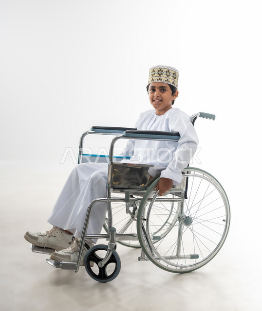 تعابير السعادة والسرور والتفاؤل, الاهتمام بالاطفال من ذوي الاحتياجات الخاصة في سلطنة عمان, بورتريه لطفل عربي خليجي عماني يرتدي الدشداشة و الكمه يجلس على كرسي متحرك ينظر الى الكاميرا بايماءات البراءة, خلفية بيضاء