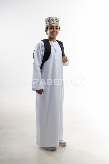 ايماءات تدل على السعادة و السرور، الاستعداد للذهاب إلى المدرسة، بورتريه لطفل عربي خليجي عماني يرتدي الدشداشة و الكمه يحمل على ظهرة حقيبة مدرسية سواء، بدء العام الدراسي الجديد في سلطنة عمان، صورة الجسم بالطول الكامل, خلفية بيضاء