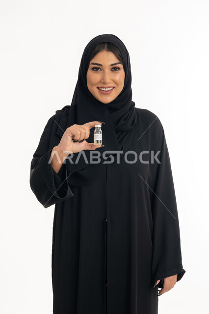 اتباع اجراءات وزارة الصحة، التطعيم ضد الفايروسات، الوعي الصحي وتجنب إنتشار العدوى، بورتريه لامرأة عربية خليجة اماراتية محجبة تحمل بيدها جرعة من لقاح كورونا (كوفيد -19 )، شابة سعودية تنظر الى الكاميرا بايماءات السرور والسعادة، خلفية بيضاء