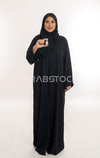 Vaccination against viruses, health awareness and avoiding the spread of infection, portrait of a veiled Emirati Gulf Arab woman holding a dose of the Corona vaccine (Covid-19), a young Saudi woman looking at the camera with gestures of pleasure and happiness, following the procedures of the Ministry of Health, a full body image, white background