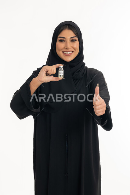 تعابير القبول والموافقة، الوعي الصحي وتجنب إنتشار العدوى، بورتريه لامرأة عربية خليجة اماراتية محجبة تحمل بيدها جرعة من لقاح كورونا (كوفيد -19 ) ترفع اصبع الابهام بايماءات الاعجاب، شابة سعودية تنظر الى الكاميرا بابتسامة، اتباع اجراءات وزارة الصحة، خلفية بيضاء