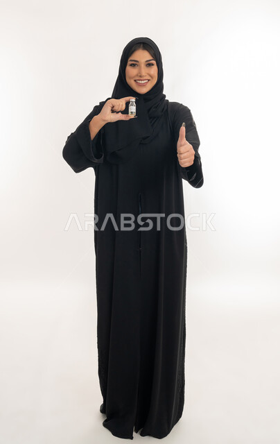 النظر الى الكاميرا بايماءات القبول، الوعي الصحي وتجنب إنتشار العدوى، بورتريه لامرأة عربية خليجة اماراتية محجبة تحمل بيدها جرعة من لقاح كورونا (كوفيد -19 )، شابة سعودية ترفع اصبع الابهام بتعابير الاعجاب، اتباع اجراءات وزارة الصحة، صورة لكامل الجسم، خلفية بيضاء