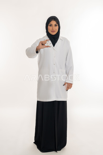 التطعيم ضد الفايروسات، اتباع اجراءات وزارة الصحة، الوعي الصحي وتجنب إنتشار العدوى، بورتريه لطبيبة عربية خليجة اماراتية محجبة ترتدي المعطف الطبي تحمل بيدها جرعة من لقاح كورونا (كوفيد -19 )، شابة سعودية تنظر الى الكاميرا بايماءات السرور، صورة لكامل الجسم، خلفية بيضاء