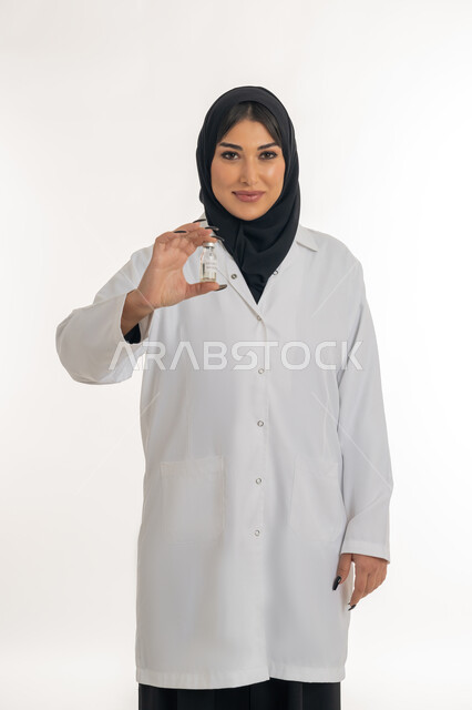 الوعي الصحي وتجنب إنتشار العدوى، التطعيم ضد الفايروسات، اتباع اجراءات وزارة الصحة، بورتريه لطبيبة عربية خليجة اماراتية محجبة ترتدي المعطف الطبي تحمل بيدها جرعة من لقاح كورونا (كوفيد -19 )، شابة سعودية تنظر الى الكاميرا بايماءات السرور، خلفية بيضاء