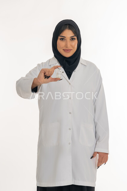 الوعي الصحي وتجنب إنتشار العدوى، التطعيم ضد الفايروسات، اتباع اجراءات وزارة الصحة، بورتريه لطبيبة عربية خليجة اماراتية محجبة ترتدي المعطف الطبي تحمل بيدها جرعة من لقاح كورونا (كوفيد -19 )، شابة سعودية تنظر الى الكاميرا بايماءات السرور، خلفية بيضاء