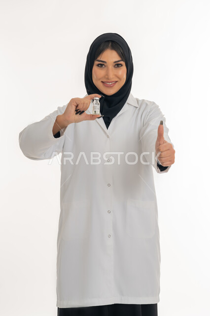 تعابير القبول والموافقة، الوعي الصحي وتجنب إنتشار العدوى، بورتريه مقرب لطبيبة عربية خليجة اماراتية محجبة ترتدي المعطف الطبي تحمل بيدها جرعة من لقاح كورونا (كوفيد - 19) ترفع اصبع الابهام بايماءات الاعجاب، شابة سعودية تقوم باتباع اجراءات الوقاية، خلفية بيضاء