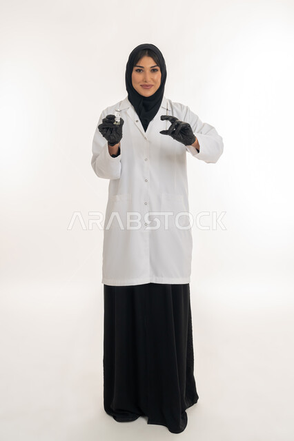 Determining volume and mass, following preventive measures, avoiding the spread of infection, portrait of a veiled Gulf Arab Emirati female doctor wearing a coat and a medical mask holding a syringe and a dose of the Corona (Covid-19) vaccine in her hand, a young Saudi woman raising her hands up to measure something, a full body image, white background