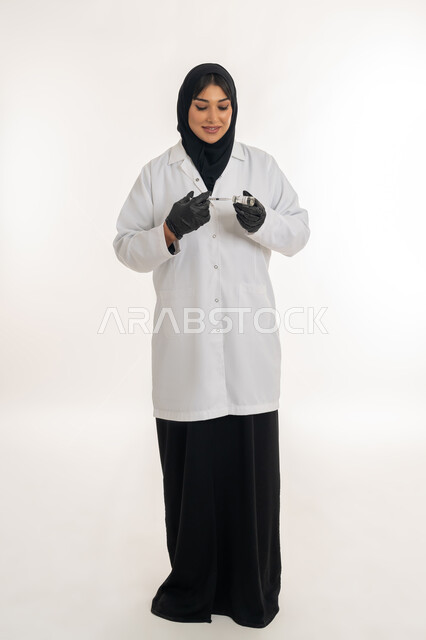 Maintaining safety and protection from diseases, health awareness and avoiding the spread of infection, portrait of a veiled Gulf Arab Emirati female doctor wearing a medical coat holding a syringe in her hand preparing the Corona vaccine (Covid-19), a young Saudi woman following preventive measures, a full body image, white background