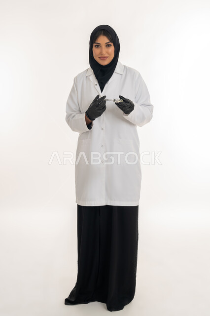 الحفاظ على السلامة والحماية من الامراض، الوعي الصحي وتجنب إنتشار العدوى، بورتريه لطبيبة عربية خليجة اماراتية محجبة ترتدي المعطف الطبي تحمل بيدها الحقنة تقوم بتجهيز لقاح كورونا (كوفيد -19 )، شابة سعودية تتبع اجراءات الوقاية، صورة لكامل الجسم، خلفية بيضاء