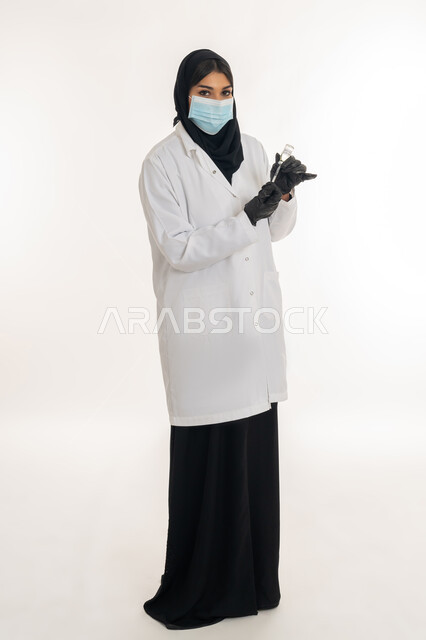 مفهوم الحماية من الامراض، الوعي الصحي وتجنب إنتشار العدوى، بورتريه من الجانب لطبيبة عربية خليجة اماراتية محجبة ترتدي المعطف الطبي تحمل بيدها الحقنة تقوم بتجهيز لقاح كورونا (كوفيد -19 )، شابة سعودية تتبع اجراءات الوقاية، صورة لكامل الجسم، خلفية بيضاء