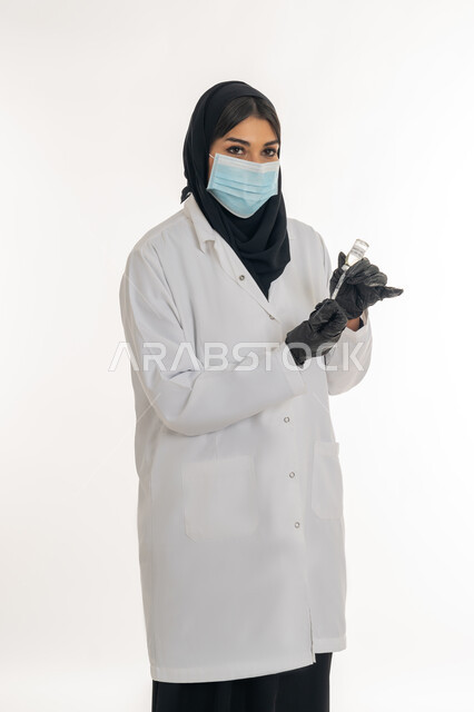 تجنب إنتشار العدوى، الحفاظ على السلامة والحماية من الامراض، مفهوم الوعي الصحي، بورتريه من الجانب لطبيبة عربية خليجة اماراتية محجبة ترتدي المعطف والكمامة الطبية تحمل بيدها الحقنة تقوم بتجهيز لقاح كورونا (كوفيد -19 )، شابة سعودية تتبع اجراءات الوقاية، خلفية بيضاء