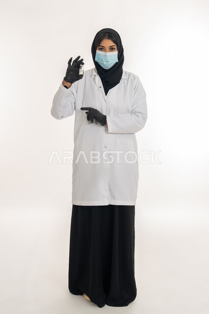 تحديد موقع شيء معين، مفهوم الوعي الصحي، تجنب إنتشار العدوى، بورتريه لطبيبة عربية خليجة اماراتية محجبة ترتدي المعطف والكمامة الطبية تحمل بيدها حقنة وجرعة من لقاح كورونا (كوفيد -19 )، شابة سعودية تشير بيدها لمكان ما، اتباع اجراءات الوقاية، صورة لكامل الجسم، خلفية بيضاء