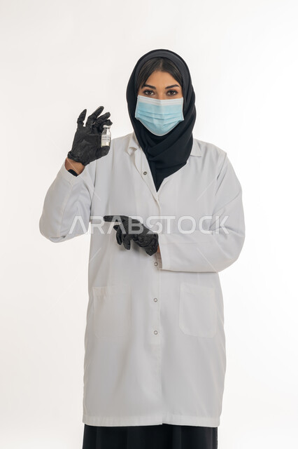 اتباع اجراءات الوقاية، تحديد موقع شيء معين، مفهوم الوعي الصحي، تجنب إنتشار العدوى، بورتريه لطبيبة عربية خليجة اماراتية محجبة ترتدي المعطف والكمامة الطبية تحمل بيدها حقنة وجرعة من لقاح كورونا (كوفيد -19 )، شابة سعودية تشير بيدها لمكان ما، خلفية بيضاء