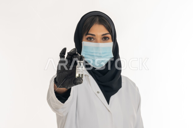 الحفاظ على السلامة والحماية من الامراض، الوعي الصحي وتجنب إنتشار العدوى، بورتريه مقرب لطبيبة عربية خليجة اماراتية محجبة ترتدي المعطف والكمامة الطبية تحمل بيدها جرعة من لقاح كورونا (كوفيد -19 )، شابة سعودية تتبع اجراءات الوقاية، خلفية بيضاء