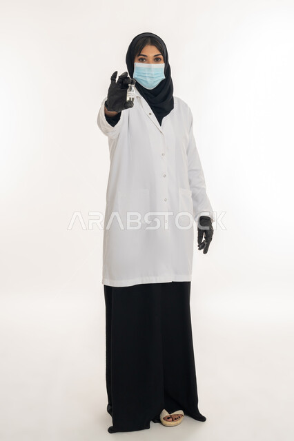 الوعي الصحي وتجنب إنتشار العدوى، الحفاظ على السلامة والحماية من الامراض، بورتريه لطبيبة عربية خليجة اماراتية محجبة ترتدي المعطف والكمامة الطبية تحمل بيدها جرعة من لقاح كورونا (كوفيد -19 )، شابة سعودية تتبع اجراءات الوقاية، صورة لكامل الجسم، خلفية بيضاء