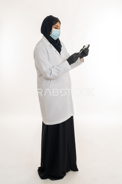 مفهوم الحماية من الامراض، الوعي الصحي وتجنب إنتشار العدوى، بورتريه من الجانب لطبيبة عربية خليجة اماراتية محجبة ترتدي المعطف الطبي تحمل بيدها الحقنة تقوم بتجهيز لقاح كورونا (كوفيد -19 )، شابة سعودية تتبع اجراءات الوقاية، صورة لكامل الجسم، خلفية بيضاء