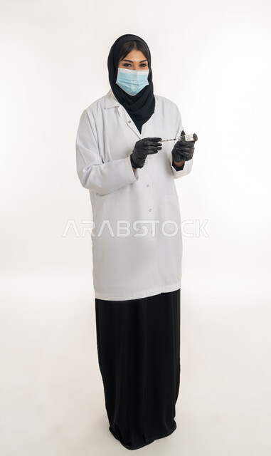 الحفاظ على السلامة والحماية من الامراض، الوعي الصحي وتجنب إنتشار العدوى، بورتريه لطبيبة عربية خليجة اماراتية محجبة ترتدي المعطف والكمامة الطبية تحمل بيدها الحقنة تقوم بتجهيز لقاح كورونا (كوفيد -19 )، شابة سعودية تتبع اجراءات الوقاية، صورة لكامل الجسم، خلفية بيضاء