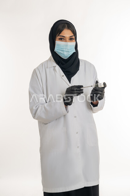 تجنب إنتشار العدوى، الحفاظ على السلامة والحماية من الامراض، مفهوم الوعي الصحي، بورتريه لطبيبة عربية خليجة اماراتية محجبة ترتدي المعطف والكمامة الطبية تحمل بيدها الحقنة تقوم بتجهيز لقاح كورونا (كوفيد -19 )، شابة سعودية تتبع اجراءات الوقاية، خلفية بيضاء
