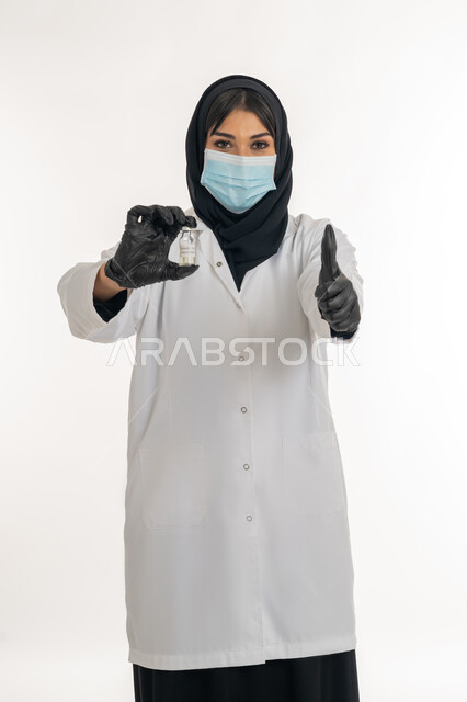 تعابير القبول والموافقة، الوعي الصحي وتجنب إنتشار العدوى، بورتريه مقرب لطبيبة عربية خليجة اماراتية محجبة ترتدي المعطف والكمامة الطبية تحمل بيده جرعة من لقاح كورونا (كوفيد - 19) ترفع اصبع الابهام بايماءات الاعجاب، شابة سعودية تقوم باتباع اجراءات الوقاية، خلفية بيضاء