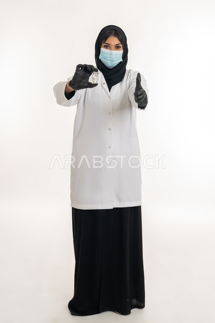 ايماءات القبول والموافقة، بورتريه لطبيبة عربية خليجة اماراتية محجبة ترتدي المعطف والكمامة الطبية تحمل بيدها جرعة من لقاح كورونا (كوفيد - 19) ترفع اصبع الابهام بتعابير الاعجاب، شابة سعودية تقوم باتباع اجراءات الوقاية، صورة لكامل الجسم، خلفية بيضاء