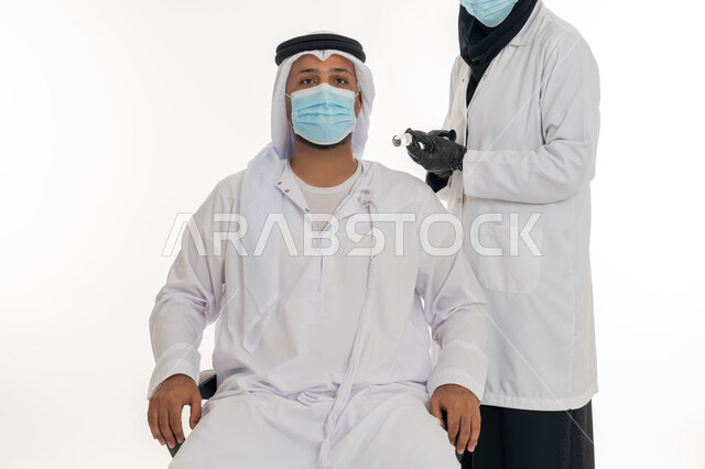 Following prevention and safety measures, health awareness and avoiding the spread of infection, receiving the Corona virus vaccination, adhering to the procedures of the Ministry of Health, portrait of an Arab Gulf Emirati man wearing a kandura, ghutra and medical mask receiving the Covid-19 vaccine from a female doctor, white background