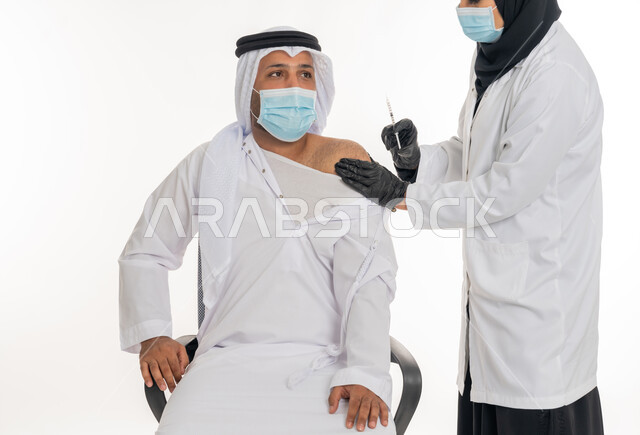 Following prevention and safety measures, health awareness and avoiding the spread of infection, receiving the Corona virus vaccination, adhering to the procedures of the Ministry of Health, portrait of an Arab Gulf Emirati man wearing a kandura, ghutra and medical mask receiving the Covid-19 vaccine from a female doctor, white background