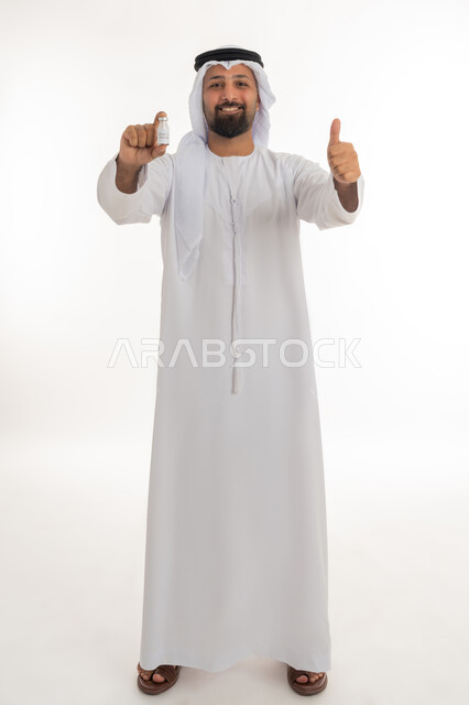 ايماءات القبول والموافقة، بورتريه لرجل عربي خليجي اماراتي يرتدي الكندورة والغترة يحمل بيده جرعة من لقاح كوفيد - 19 يرفع اصبع الابهام بتعابير الاعجاب والرضى، تلقي التطعيم ضد فايروس كورونا، اتباع اجراءات وزارة الصحة، صورة لكامل الجسم، خلفية بيضاء