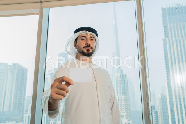 الوقوف باستقامة والنظر الى الكاميرا بايماءات السرور والسعادة، موك أب أبيض مستطيل فارغ، عرض بطاقة تعريفية، صورة مقربة لرجل عربي خليجي اماراتي يرتدي الكندورة والغترة البيضاء يحمل بيده هوية شخصية، بيئة العمل المريحة في شركات الامارات